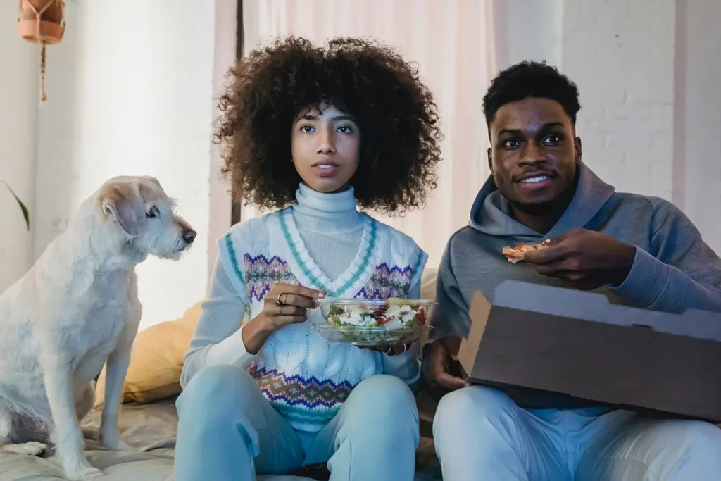 man and woman eating on a couch while a dog is watching them