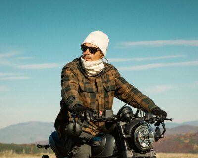Мъж със слънчеви очила на мотоциклет, с шапка Nomad Heat и шал тип гейтър за врата, с небе на заден план.